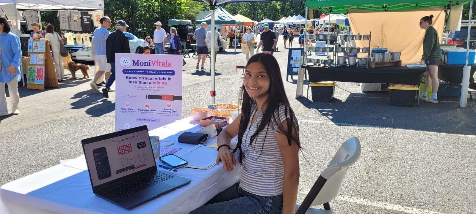 MoniVitals at a farmers market: a laptop showing how pixels become a pulse, next to the screening board